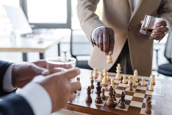 Vue recadrée d'un homme d'affaires afro-américain avec un verre de whisky jouant aux échecs avec un collègue flou — Photo de stock