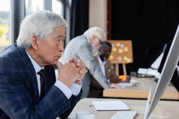 Senior asiatischer Geschäftsmann schaut auf Computermonitor in der Nähe verschwommener multiethnischer Kollegen — Stockfoto