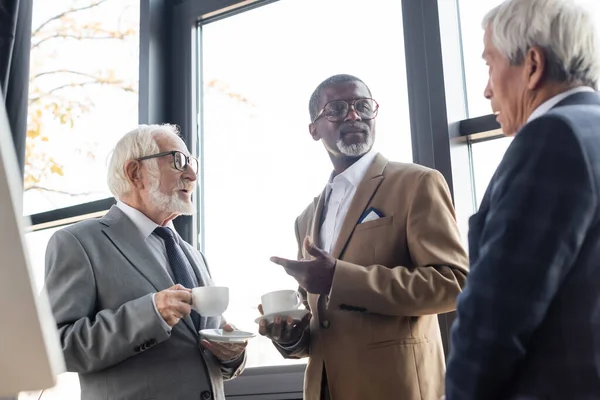 Uomo d'affari afroamericano che punta con mano mentre parla con colleghi anziani durante la pausa caffè — Foto stock