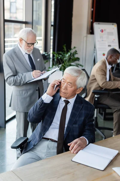 Senior asian businessman talking on smartphone near empty notebook and interracial colleagues — Stock Photo