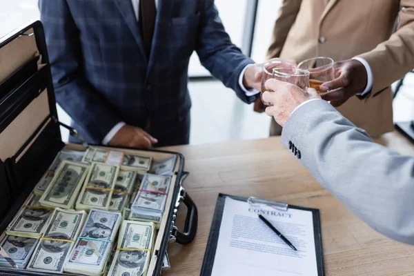 Vista cortada de parceiros de negócios clinking copos de uísque perto de contrato e pasta com dinheiro — Fotografia de Stock