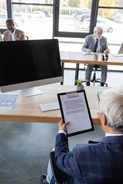 Empresário sênior contrato de leitura enquanto sentado perto do monitor do computador com tela em branco — Fotografia de Stock