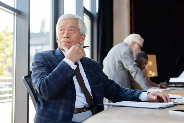 Senior asiatique homme d'affaires regardant loin tout en travaillant près flous collègues multiethniques — Stock Photo