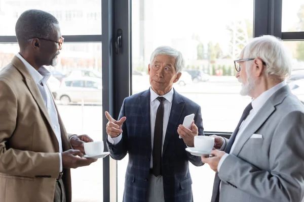 Senior asiatico uomo d'affari con smartphone punta con dito mentre parla con interrazziale colleghi su caffè pausa — Foto stock