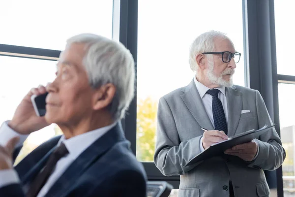 Verschwommener asiatischer Geschäftsmann spricht auf Smartphone neben Seniorenkollege und schreibt auf Klemmbrett — Stockfoto