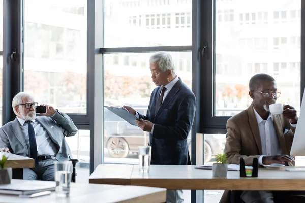 Senior asiático hombre de negocios de pie con portapapeles cerca multiétnico colegas trabajando en oficina - foto de stock