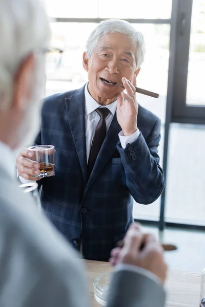 Vieux asiatique homme d'affaires avec cigare et verre de whisky souriant près flou collègue — Photo de stock