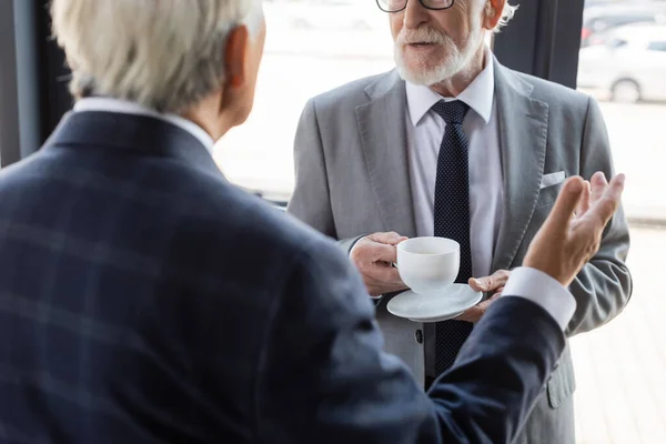 Vista recortada de hombre de negocios senior con taza de café cerca de colega borrosa señalando con la mano - foto de stock