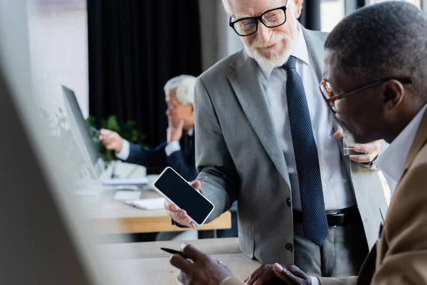 Senior businessman showing smartphone with blank screen to african american colleague in office — Stock Photo