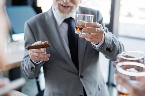 Vista recortada del anciano hombre de negocios sosteniendo cigarro y vaso de whisky cerca de colegas borrosos - foto de stock