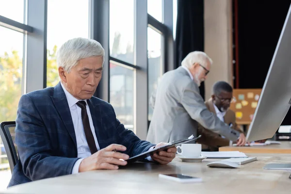 Senior asiático hombre de negocios trabajando con documento cerca borrosa interracial colegas - foto de stock