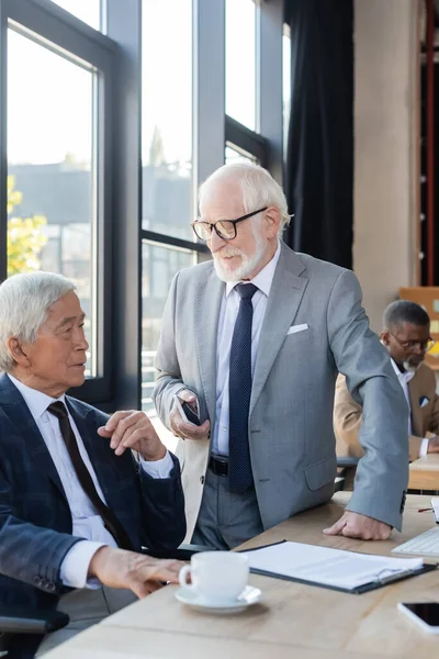 Senior businessman with mobile phone talking to asian colleague near blurred contract on desk — Stock Photo
