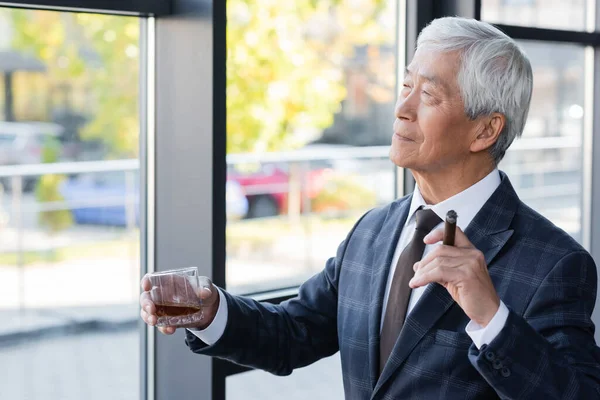 Homme d'affaires asiatique âgé regardant loin tout en se tenant près de la fenêtre avec cigare et verre de whisky — Photo de stock