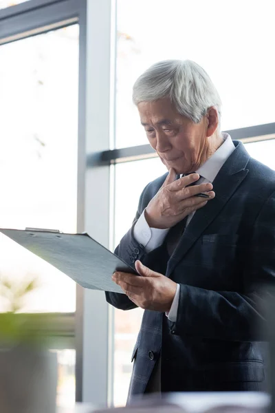 Leitender asiatischer Geschäftsmann denkt beim Anblick des Klemmbretts im Büro — Stockfoto