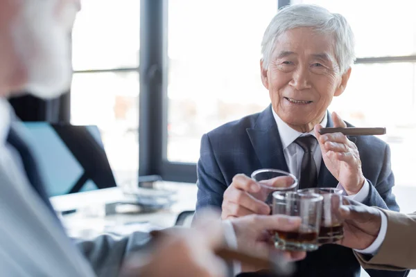 Senior asian businessman with cigar clinking glasses with blurred multiethnic business partners in office — Stock Photo