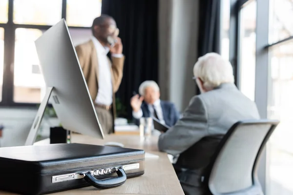 Selective focus of briefcase on desk near multiethnic senior colleagues working on blurred background — Stock Photo
