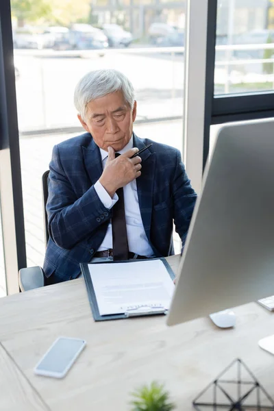 Pensativo asiático hombre de negocios trabajando con contrato cerca de computadora en lugar de trabajo - foto de stock