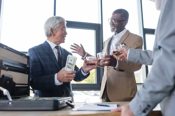 Feliz asiático homem de negócios com notas de dólar perto de parceiros de negócios multiétnicos com copos de uísque — Fotografia de Stock