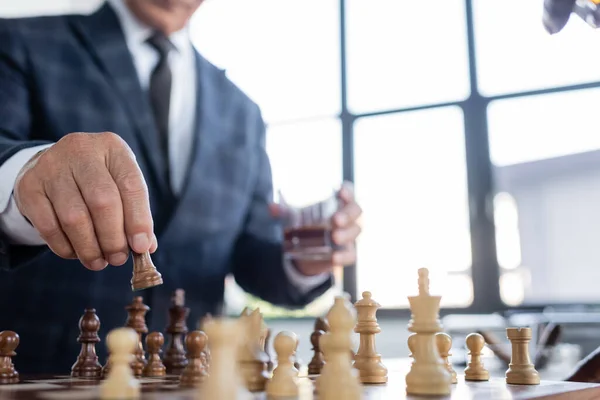 Vue partielle d'un homme d'affaires flou avec un verre de whisky jouant aux échecs au bureau — Photo de stock