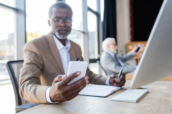 Blurred african american businessman looking at smartphone while working near blurred colleague — Stock Photo