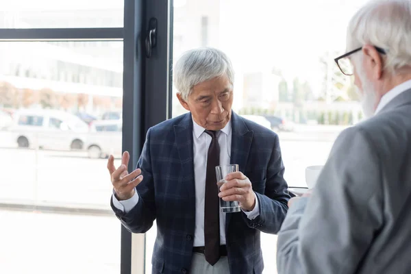 Anziano asiatico uomo d'affari holding bicchiere d'acqua durante discussione con blurred collega — Foto stock
