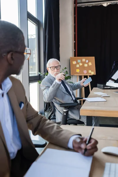 Elderly businessman with cellphone talking to african american colleague working on blurred foreground — Stock Photo
