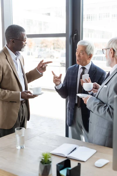 Senior multiethnic business partners talking during coffee break near blank notebook on desk — Stock Photo