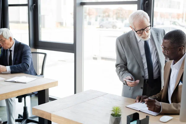 Senior businessman mostrando cellulare per africano americano business partner vicino di lavoro asiatico collega — Foto stock