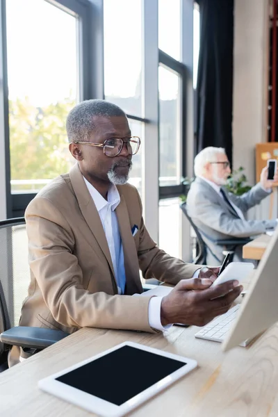 Senior afrikanisch-amerikanischer Geschäftsmann mit Brille schaut auf Smartphone, während er in der Nähe eines verschwommenen Kollegen arbeitet — Stockfoto
