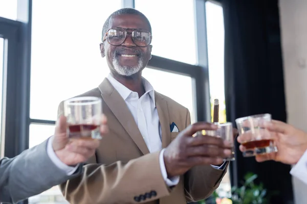 Cheerful african american businessman looking at camera near blurred colleagues with glasses of whiskey — Stock Photo