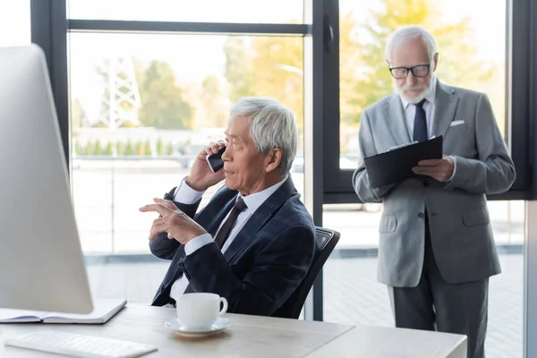 Sênior asiático empresário falando no smartphone perto de computador e colega com prancheta — Fotografia de Stock