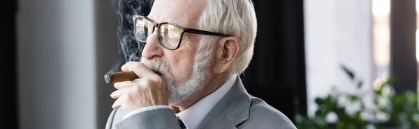 Homme d'affaires senior dans les lunettes regardant loin tout en fumant cigare dans le bureau, bannière — Photo de stock