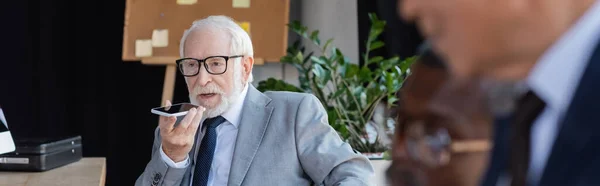 Elderly businessman in eyeglasses sending voice message on cellphone near blurred colleague, banner — Stock Photo