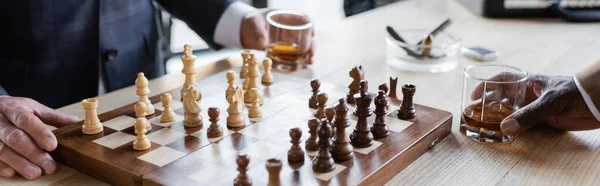 Vista recortada de los socios de negocios con vasos de whisky jugando ajedrez en la oficina, pancarta - foto de stock