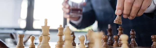 Cropped view of businessman with blurred glass of whiskey playing chess in office, banner — Stock Photo