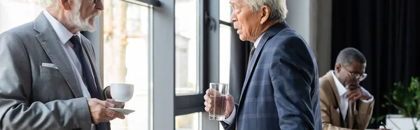 Senior businessmen with drinks talking while african american colleague working on blurred background, banner — Stock Photo