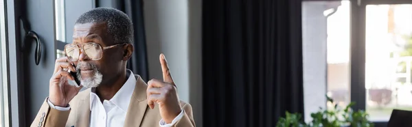 Hombre de negocios afroamericano senior mostrando gesto de idea mientras habla por teléfono móvil en la oficina, pancarta - foto de stock
