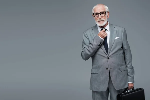 Anciano hombre de negocios con maletín ajustando corbata mientras mira la cámara aislada en gris - foto de stock