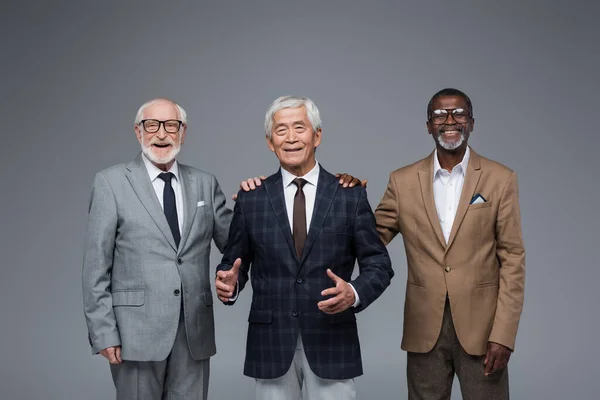 Smiling multiethnic businessmen touching shoulders of senior asian colleague isolated on grey — Stock Photo