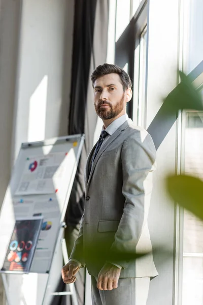 Hombre de negocios en traje mirando la cámara cerca de rotafolio borroso en la oficina - foto de stock