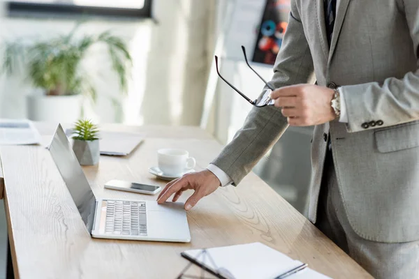 Vue recadrée de l'homme d'affaires tenant des lunettes et utilisant un ordinateur portable près du café dans le bureau — Photo de stock