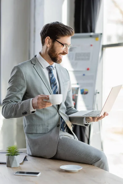 Un hombre de negocios sonriente sosteniendo una taza de café y un portátil en la oficina - foto de stock