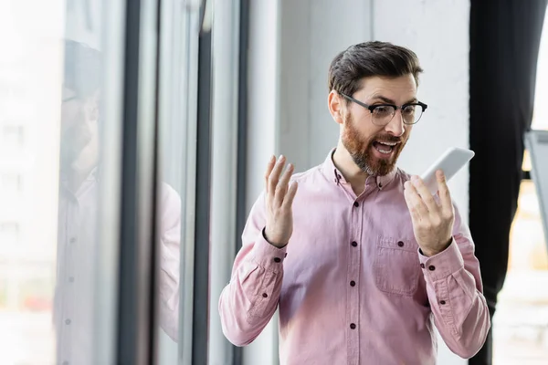 Angry businessman in eyeglasses recording voice message on smartphone in office — Stock Photo