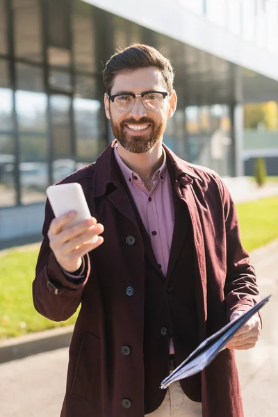 Uomo d'affari sorridente in trench che tiene smartphone e cartella di carta all'aperto — Foto stock