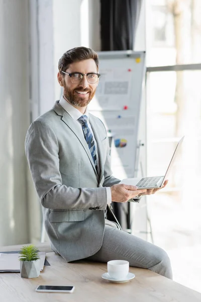 Smiling businessman in suit holding laptop near smartphone and coffee in office — Stock Photo