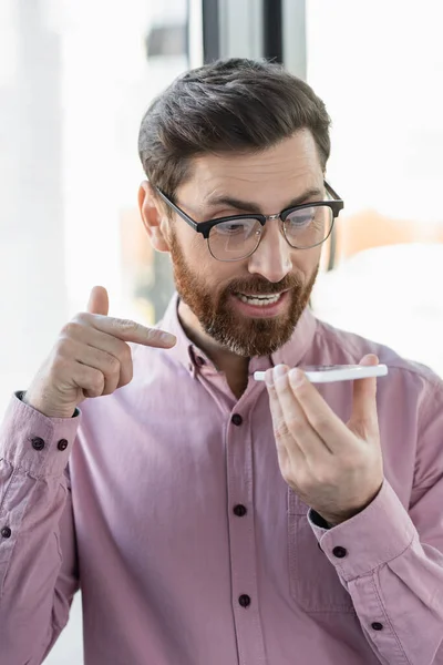 Businessman in eyeglasses recording voice message on smartphone and pointing with finger in office — Stock Photo