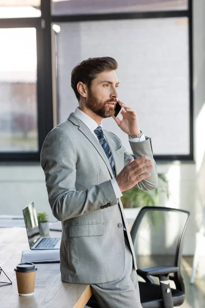 Businessman talking on mobile phone near paper cup and laptop in office — Stock Photo