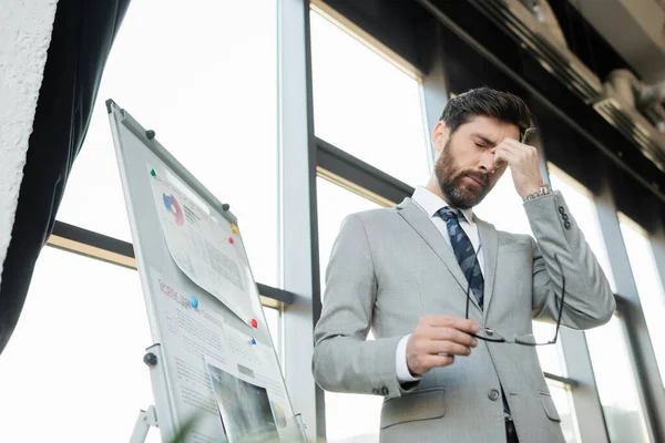 Low angle view of tired businessman holding eyeglasses near flip chart in office — Stock Photo