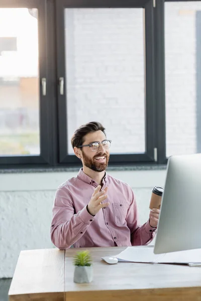 Cheerful businessman holding takeaway coffee during video call on computer in office — Stock Photo