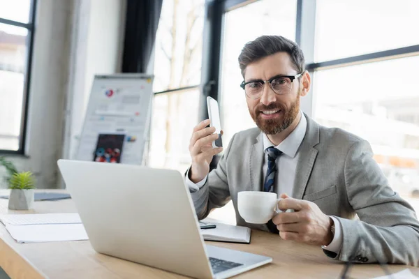 Uomo d'affari sorridente con smartphone che tiene il caffè e guardando la fotocamera vicino al laptop — Foto stock